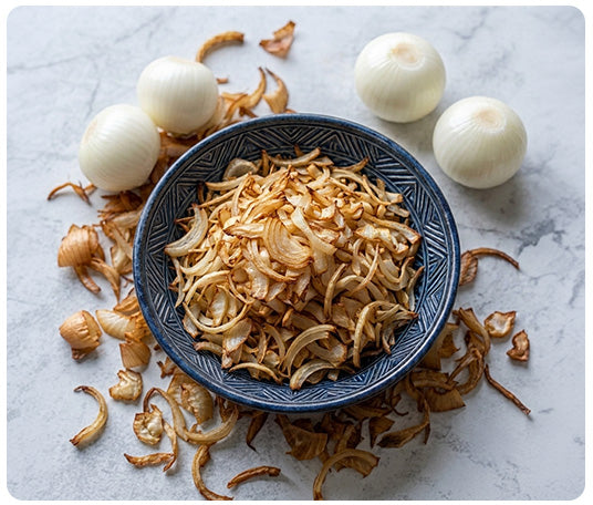 Bowl of crispy fried onions surrounded by fresh onions and toasted onion slices, ideal for cooking, garnishing, and adding rich onion flavor to everyday recipes.
